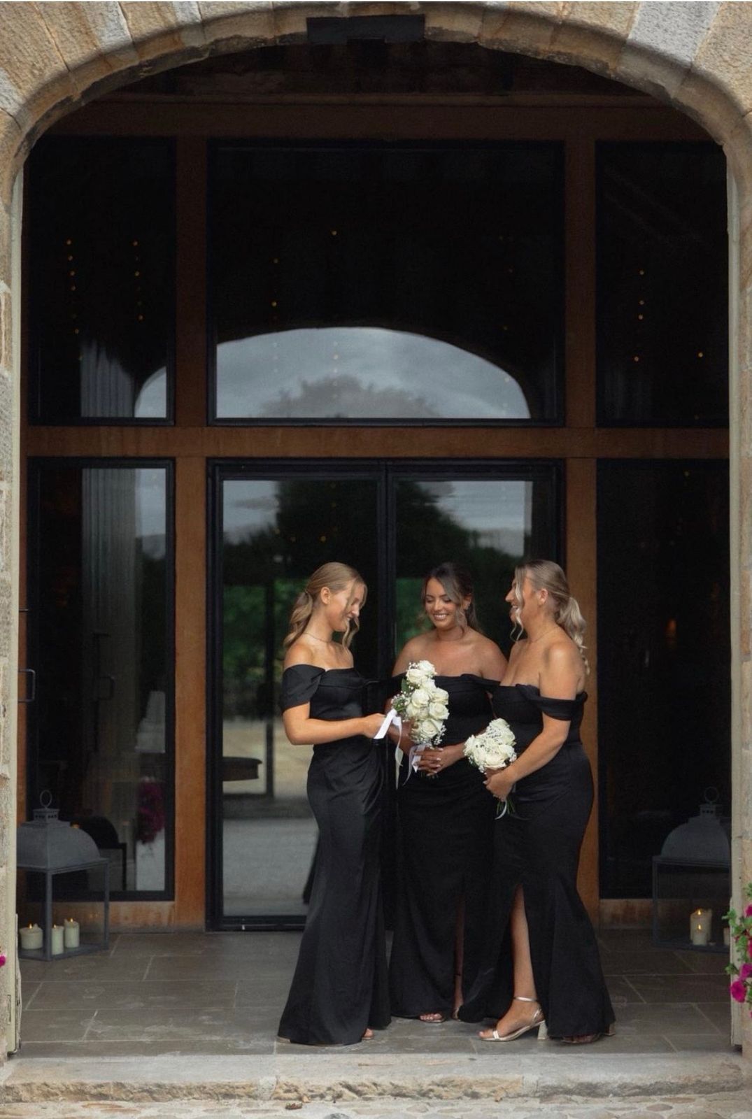 Three bridesmaids in black dresses holding white bouquets beneath a stone archway